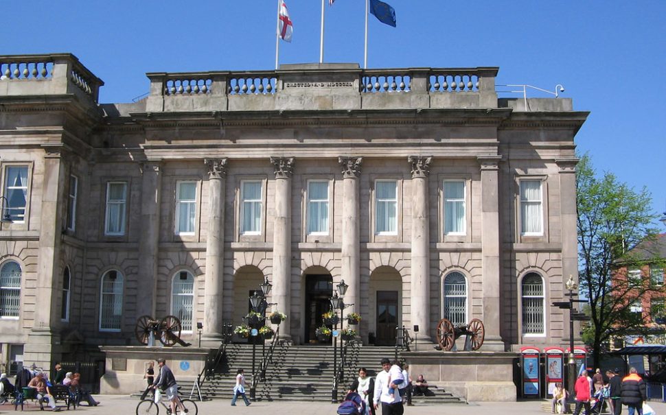 A landscape image of Ashton-under-Lyne Town Hall against a blue sky. On top of the building, three flags are flying: the Union Jack in the centre, the English flag to the left and the European Union flag to the right.