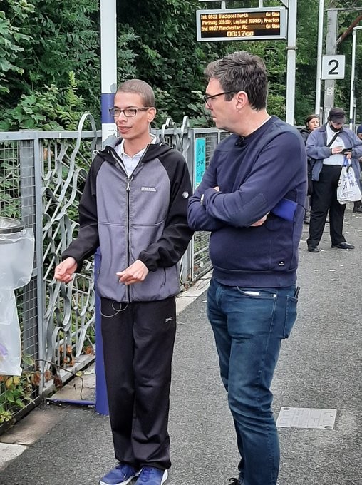 A photo of Nathaniel Yates standing next to Andy Burnham at a train station. Burnham has his arms folded and is looking at Yates who appears to be explaining something.