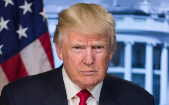 Donald Trump in the foreground, wearing a dark suit and red tie, behind him the American flag and part of the White House is visible. backgroun