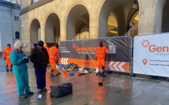 The front of manchester city library, two people one dressed in teal and one in black talk to the left of the image. In the background two sanitation workers clear rubbish from a stone floor.