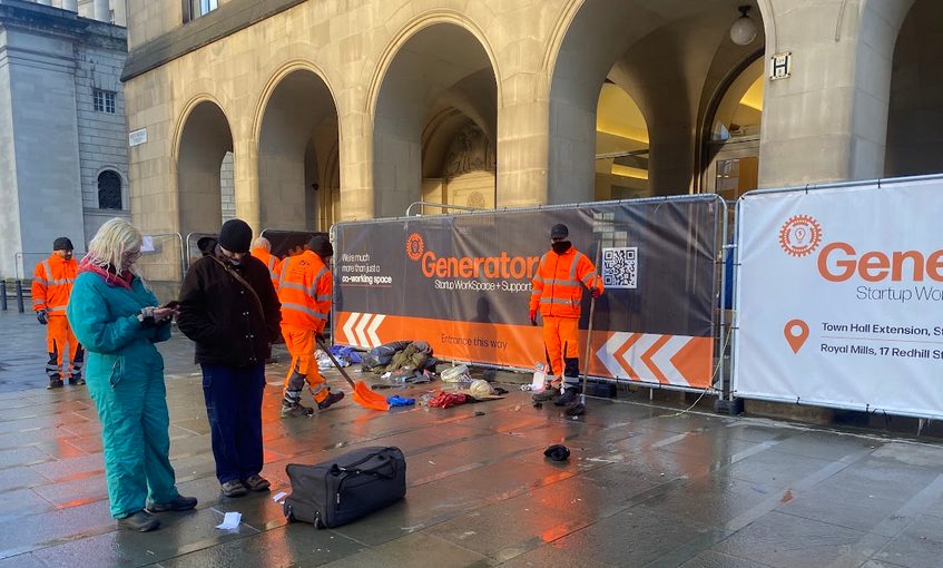 The front of manchester city library, two people one dressed in teal and one in black talk to the left of the image. In the background two sanitation workers clear rubbish from a stone floor.